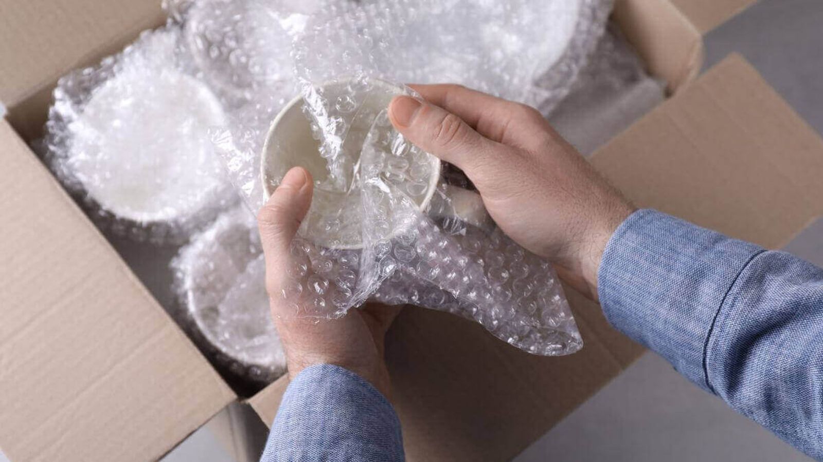 Manos envuelven un tazón de cerámica en poliburbuja sobre una caja de cartón con más piezas ya envueltas