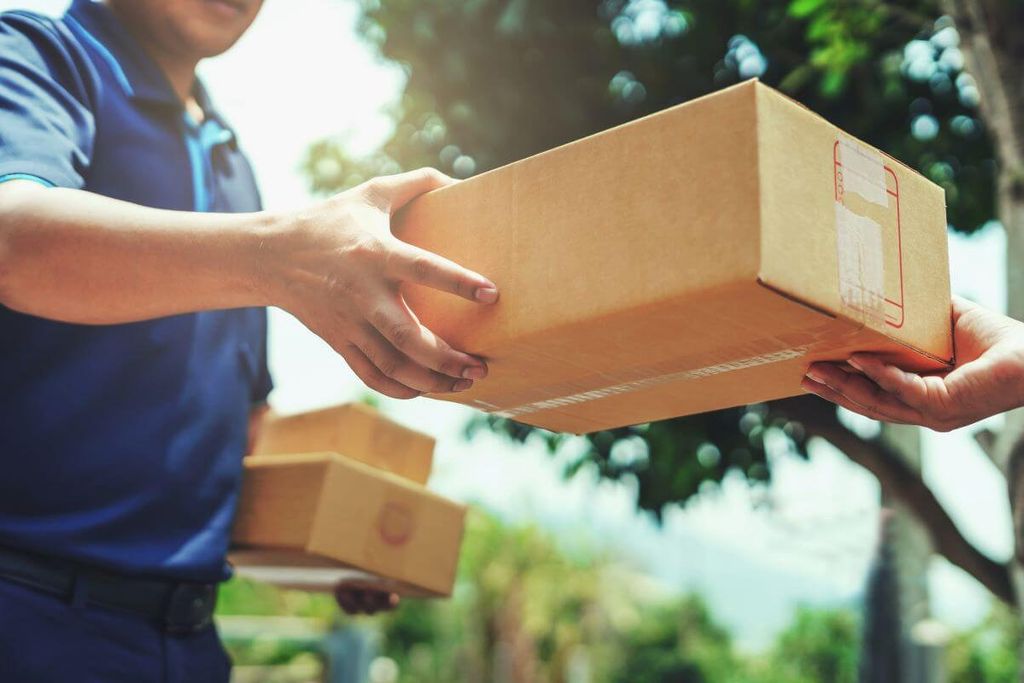 Repartidor con playera azul entrega una caja de cartón con etiqueta de envío en la mano de un cliente al aire libre