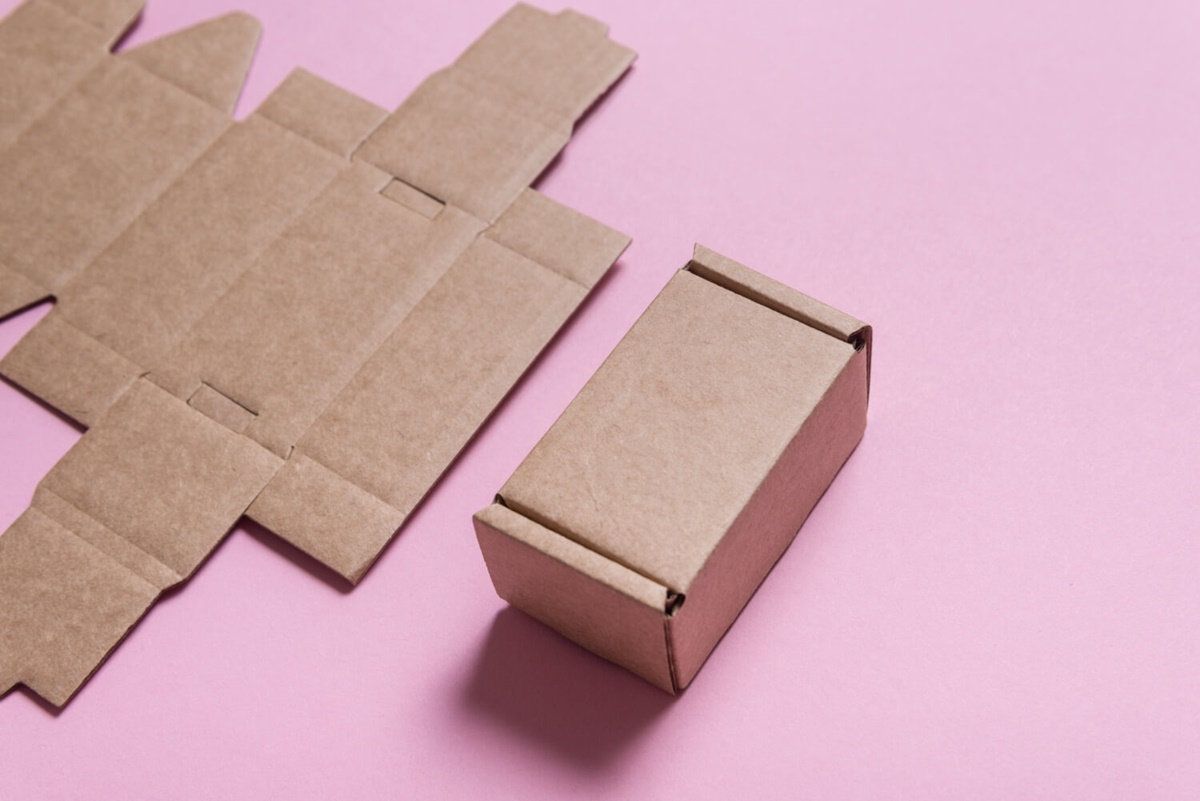 Caja troquelada de cartón kraft armada junto a su plantilla plana desplegada, sobre fondo rosa