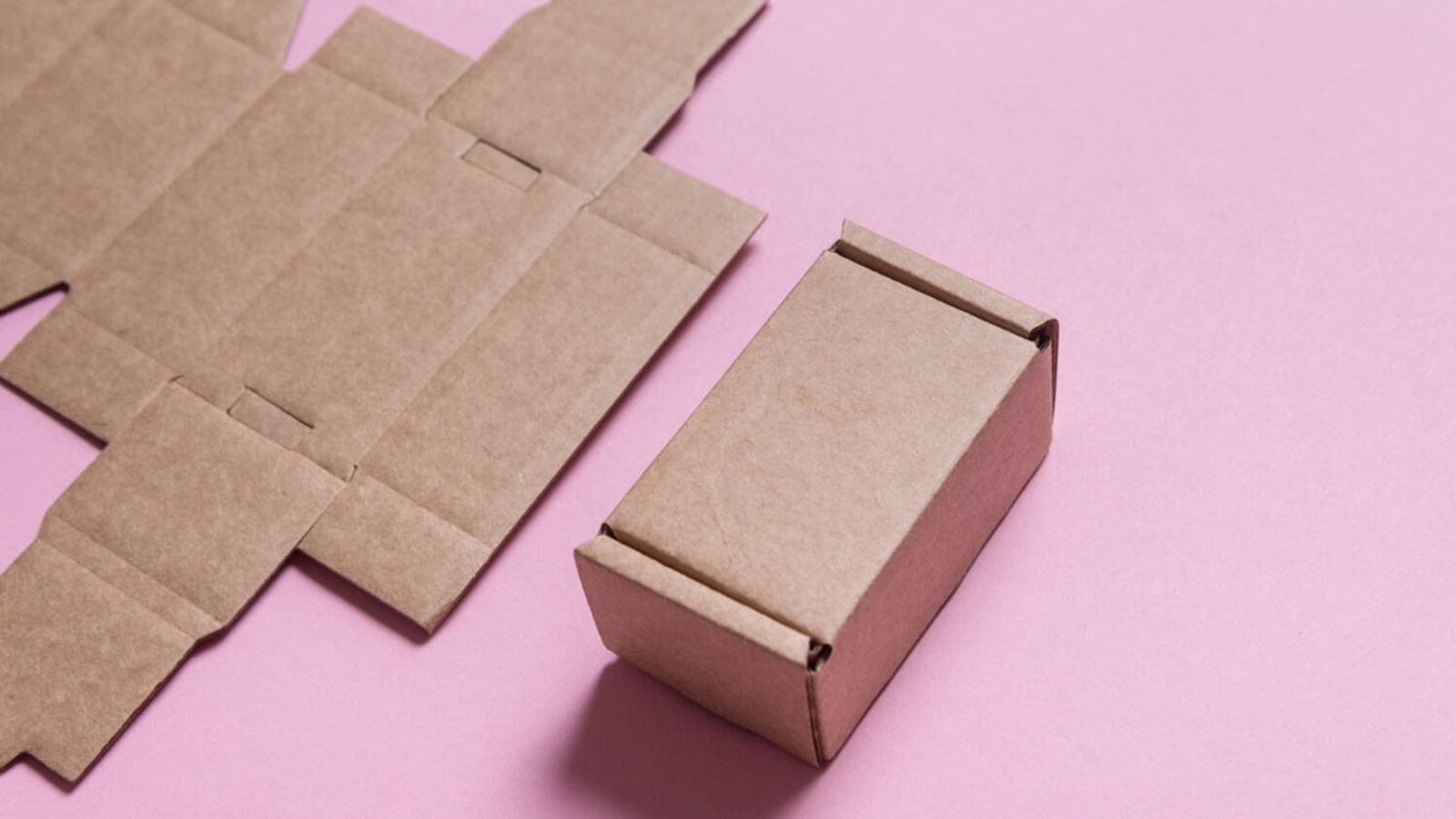 Caja troquelada de cartón kraft armada junto a su plantilla plana desplegada, sobre fondo rosa