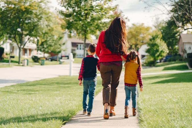 Mujer camina por la banqueta de un barrio tomada de la mano de dos niños, vistos de espaldas