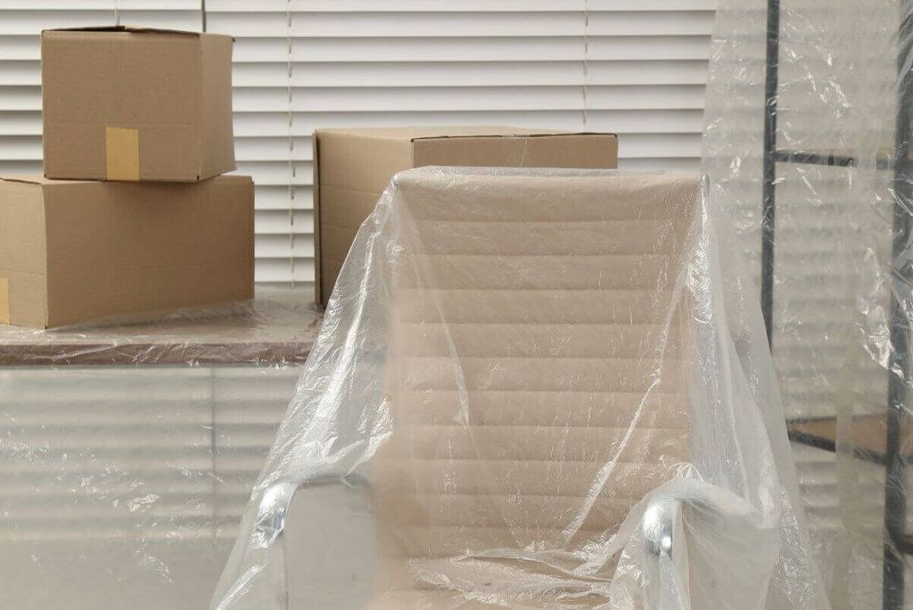 Silla de oficina cubierta con plástico frente a una persiana, con cajas de cartón al fondo