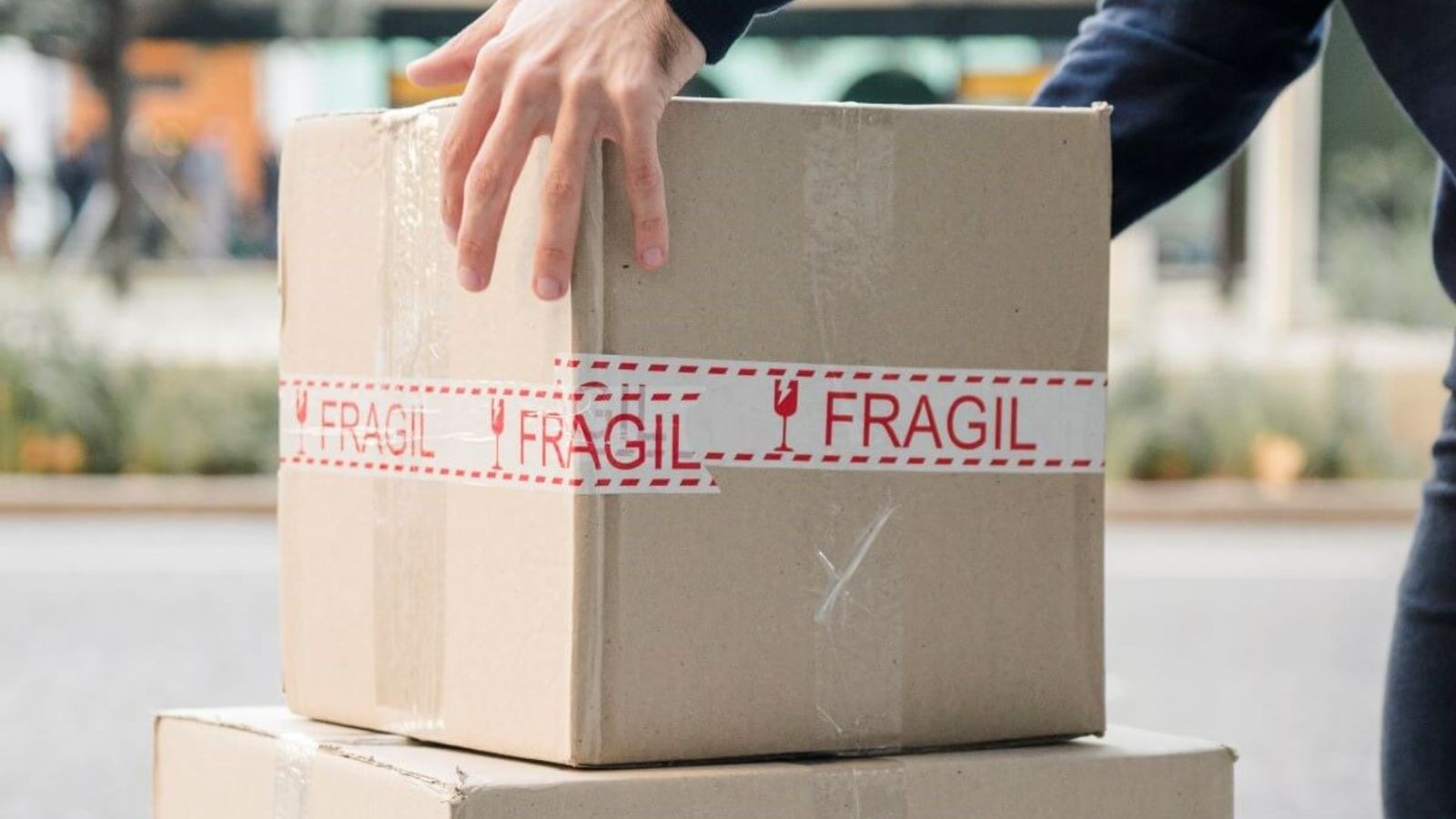 Mano sobre una caja de cartón sellada con cinta impresa FRÁGIL, apilada sobre otra caja en la calle