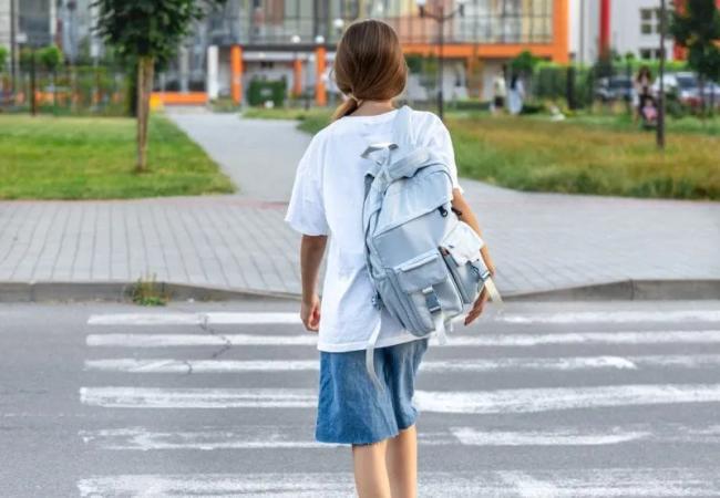 Niña con mochila cruzando una calle por el paso de cebra frente a un parque