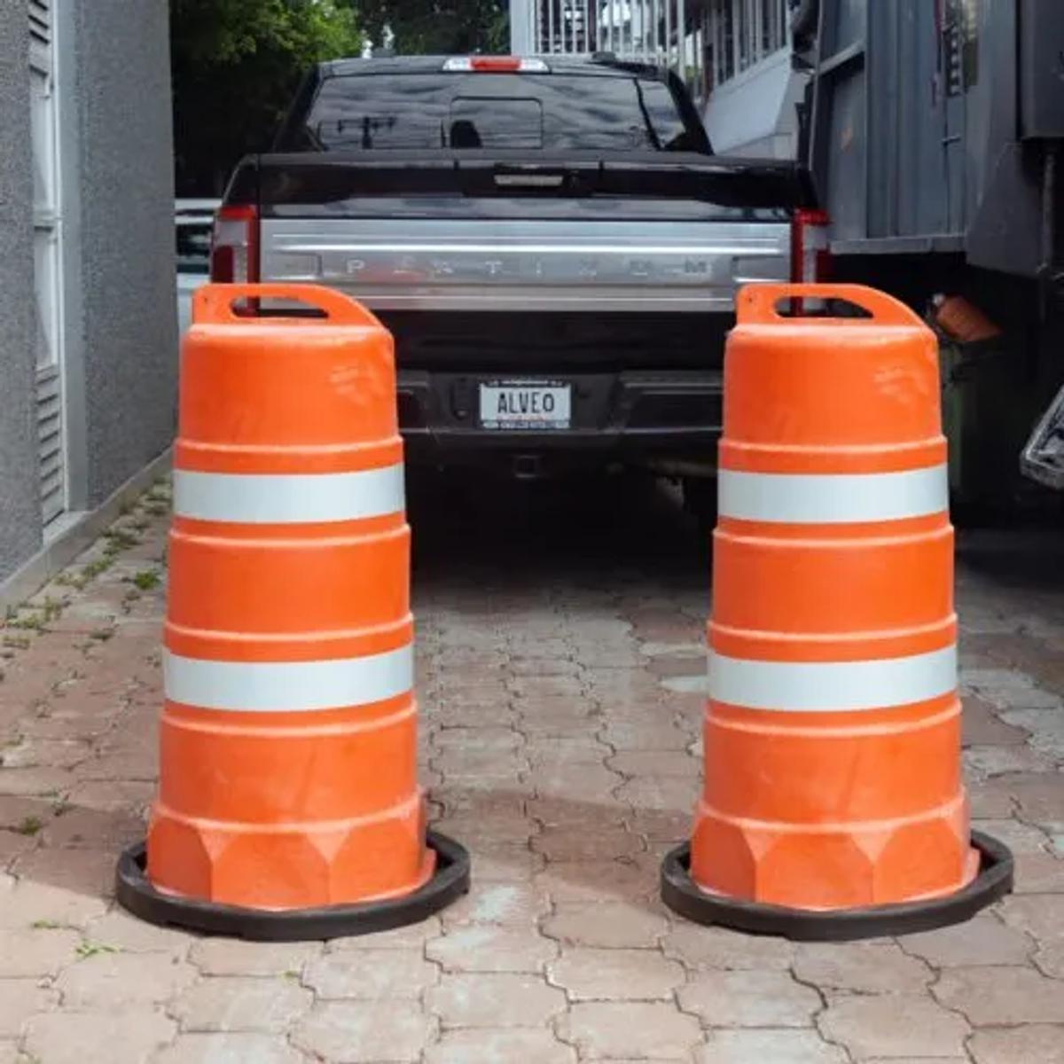 Dos trafitambos naranjas con dos bandas reflejantes sobre bases negras en una entrada de adoquín, frente a una camioneta