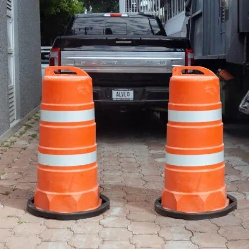 Dos trafitambos naranjas con dos bandas reflejantes sobre bases negras en una entrada de adoquín, frente a una camioneta