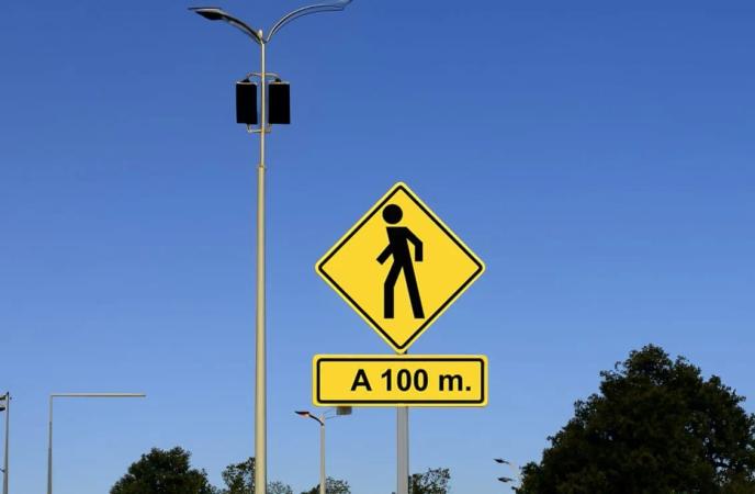 Señal preventiva amarilla de cruce de peatones con placa A 100 m, junto a un poste de alumbrado y bajo cielo azul