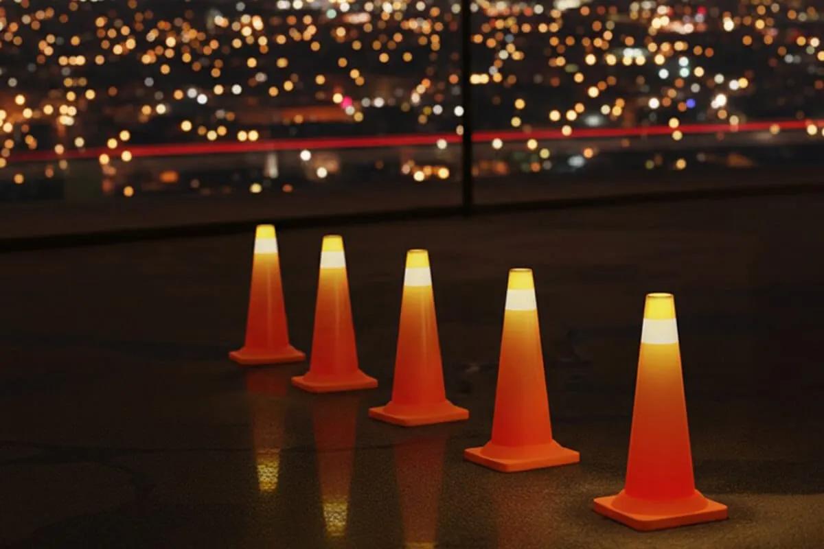 Cinco conos naranjas con luz en la punta sobre un piso oscuro, con las luces de la ciudad al fondo