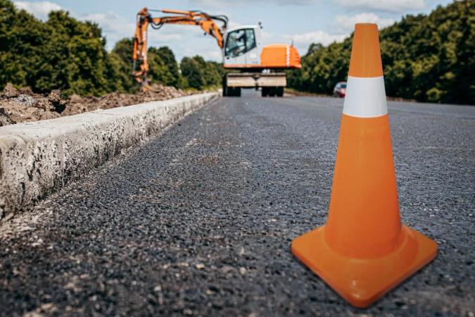Cono vial naranja con franja reflejante sobre asfalto nuevo, con una excavadora trabajando al fondo