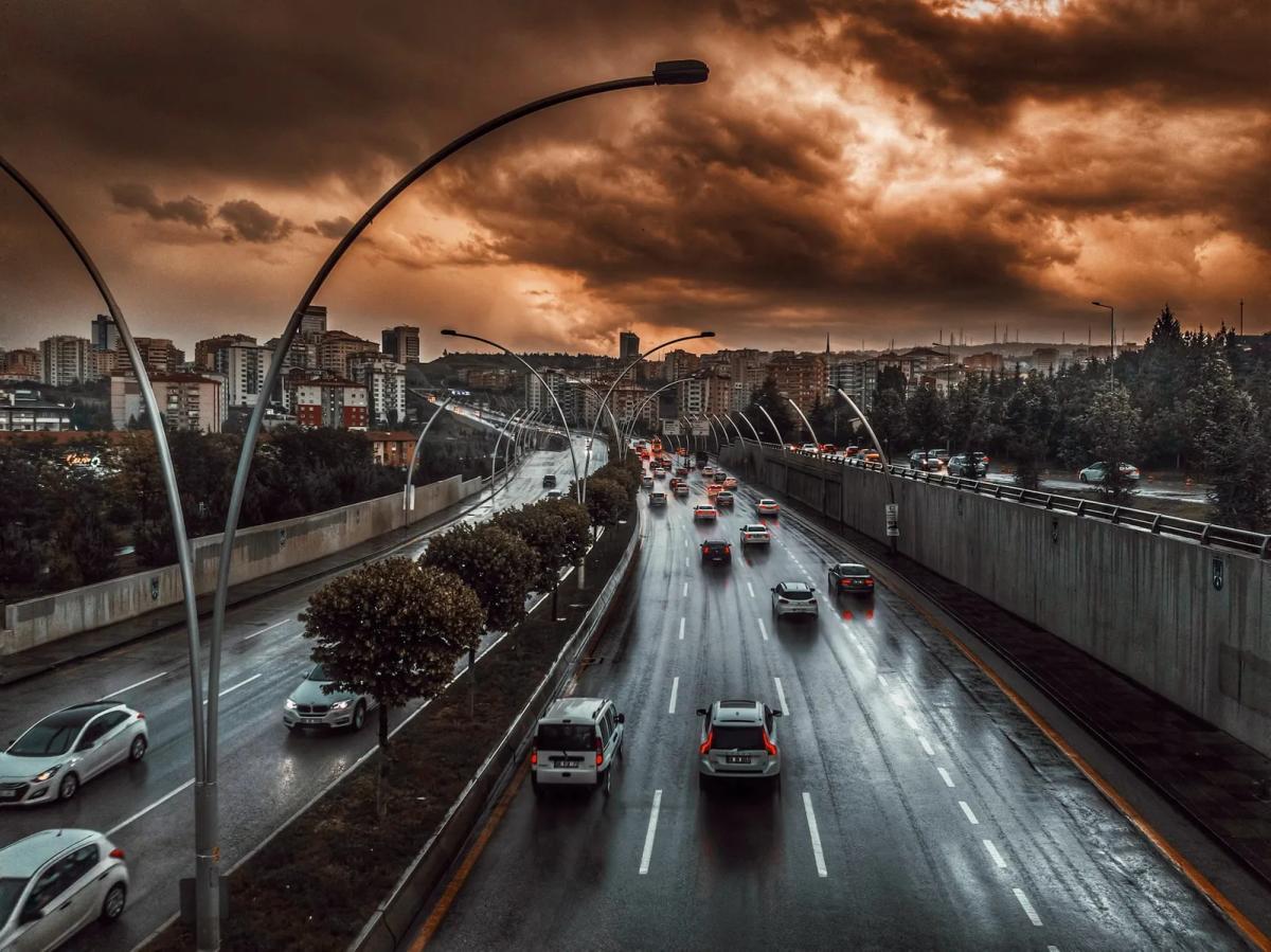 Vía rápida urbana mojada por la lluvia al atardecer, con autos de luces encendidas y nubes de tormenta