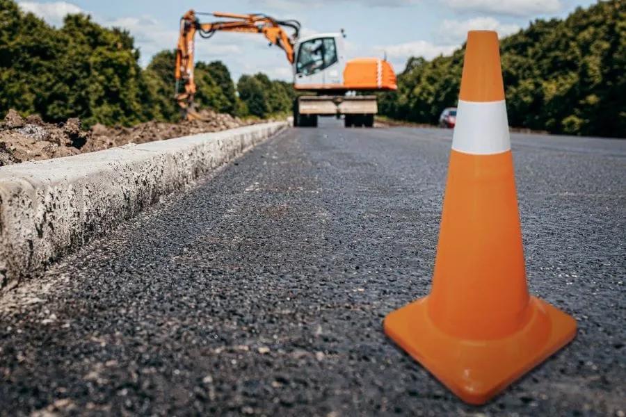 Cono de tránsito naranja con banda reflejante blanca sobre asfalto nuevo y una excavadora naranja al fondo de la obra