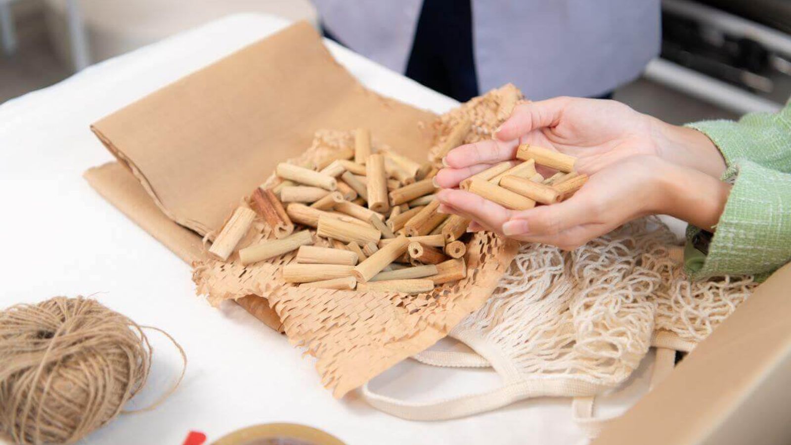 Manos sostienen pequeños tubos de cartón de relleno sobre papel kraft de panal, con hilo de yute y bolsa de malla al lado