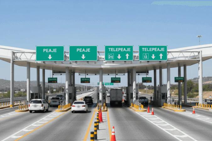 Caseta de cobro con letreros verdes de peaje y telepeaje sobre los carriles, autos en fila y conos naranjas entre carriles
