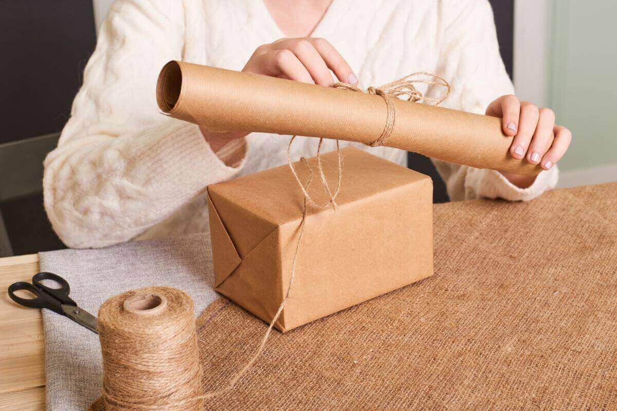 Persona sostiene un rollo de papel kraft sobre una caja envuelta en kraft y atada con hilo de yute, con tijeras en la mesa