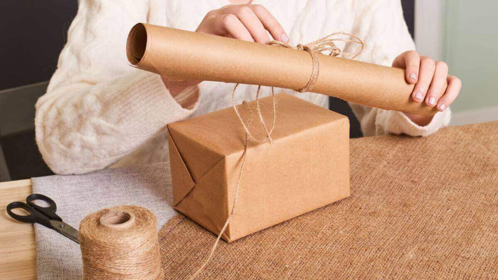 Persona sostiene un rollo de papel kraft sobre una caja envuelta en kraft y atada con hilo de yute, con tijeras en la mesa