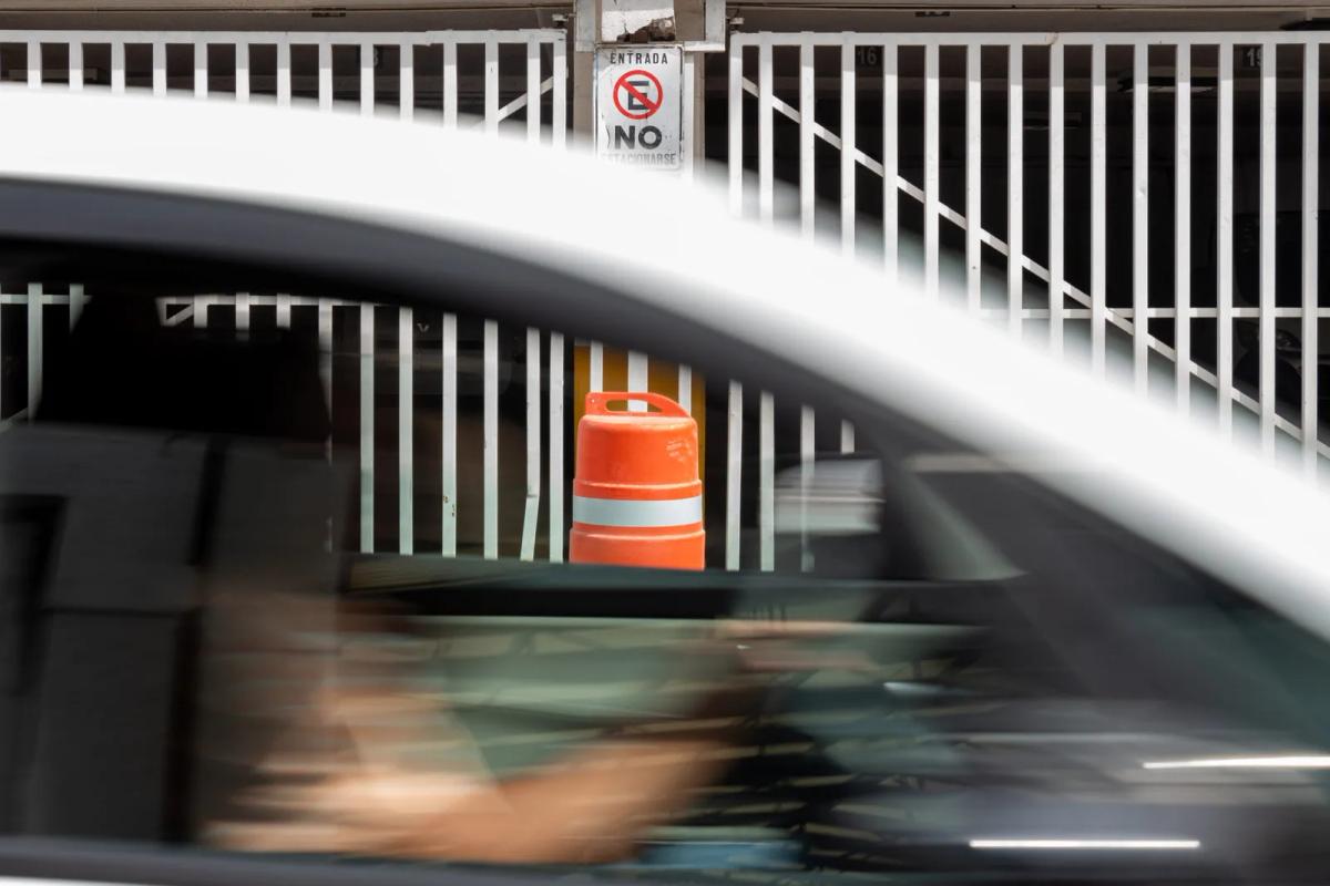Trafitambo naranja con banda reflejante frente a un portón blanco, visto tras la ventana de un auto blanco en movimiento