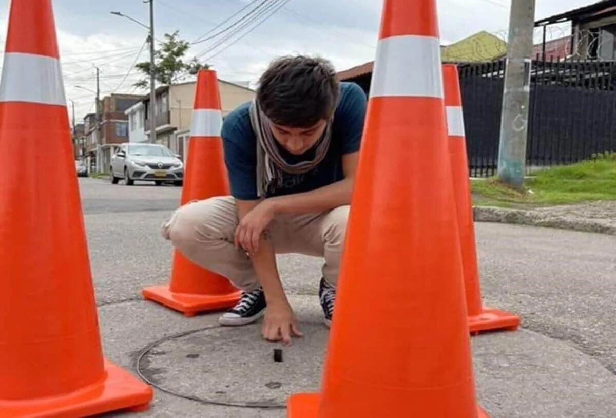 Joven en cuclillas junto a una tapa de registro en la calle, rodeado de cuatro conos viales naranjas con franja reflejante
