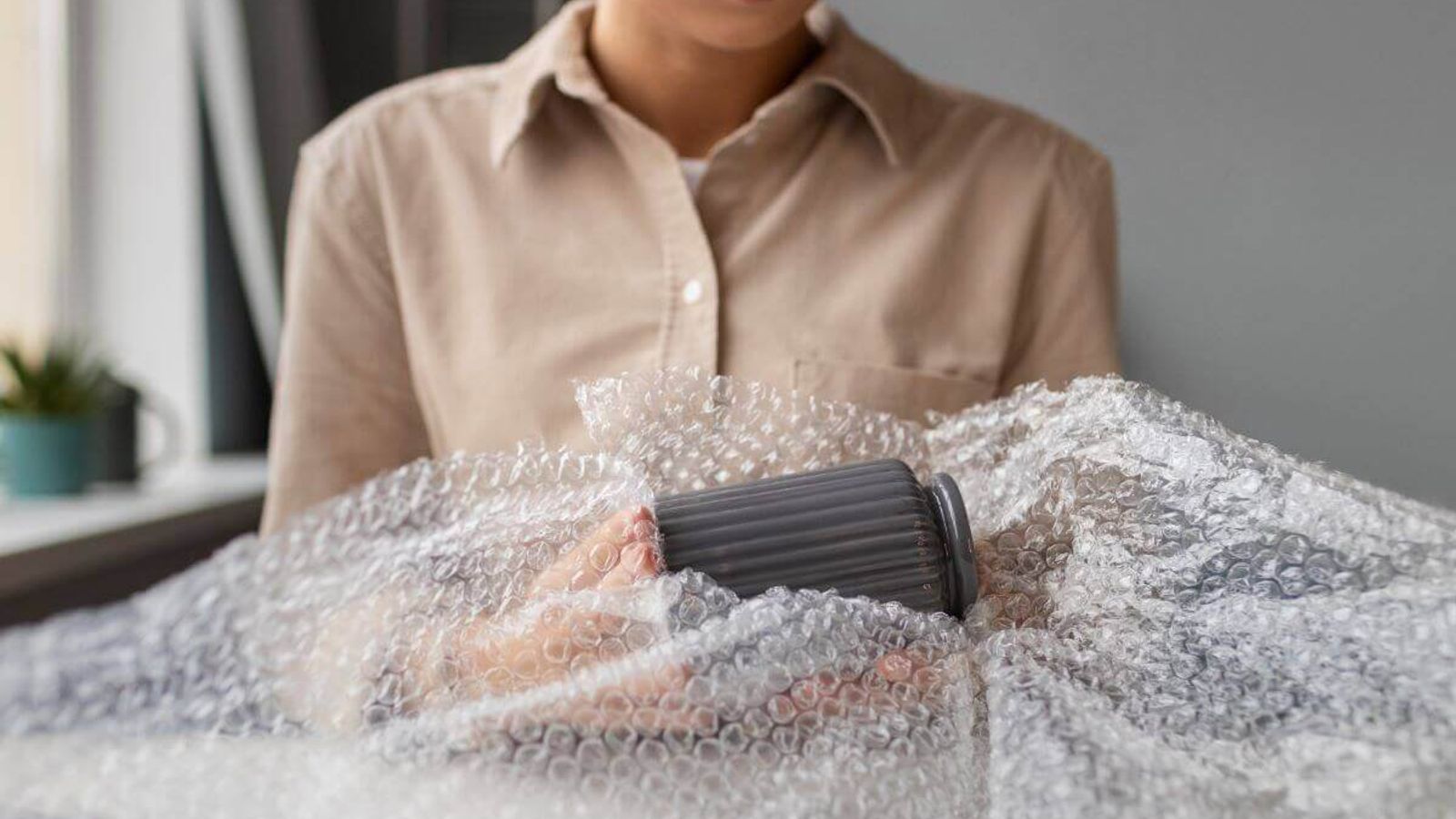 Persona con camisa beige envuelve una taza gris con poliburbuja sobre una mesa