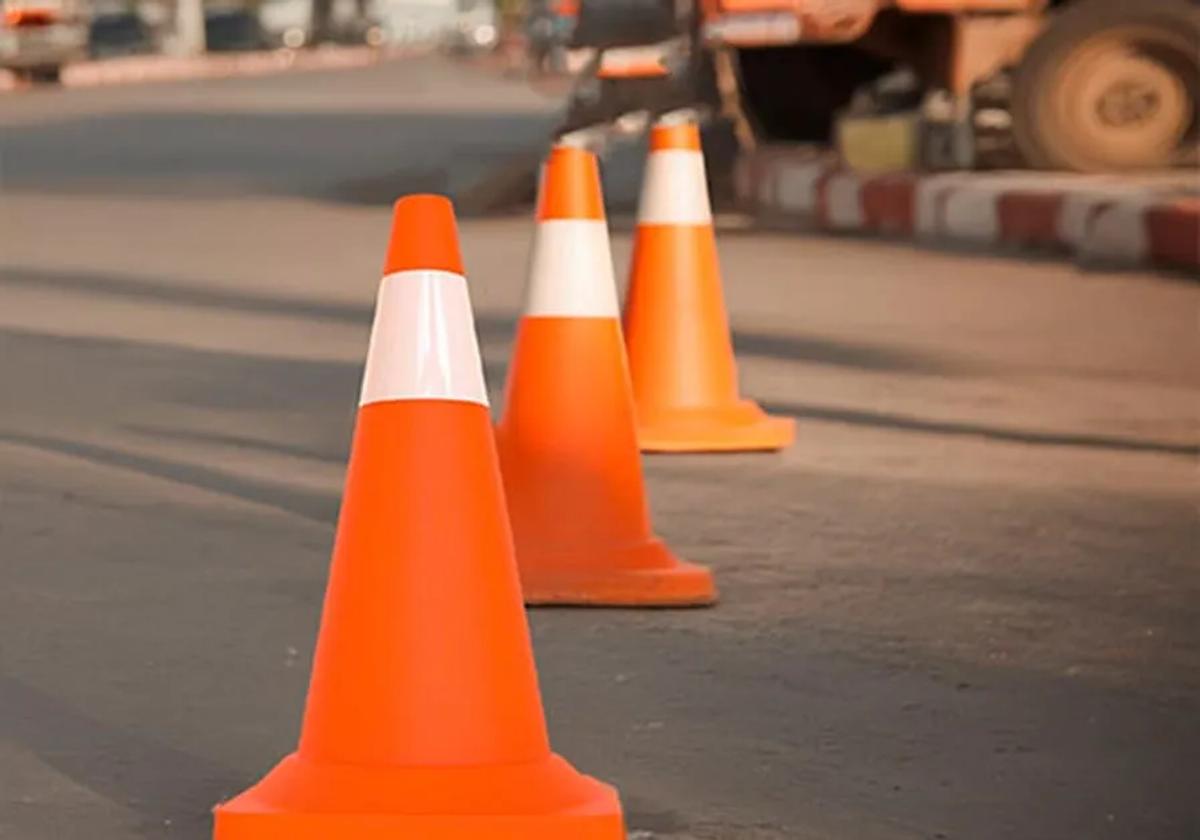 Conos viales naranja con franja reflejante alineados en una calle, con maquinaria al fondo