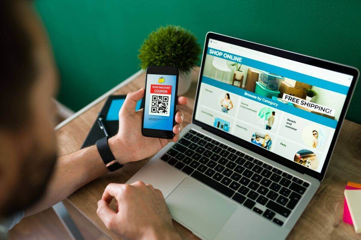 Hombre sostiene un celular con un cupón QR frente a una laptop que muestra una tienda en línea