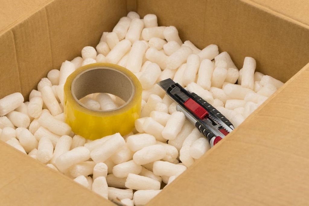 Caja de cartón llena de cacahuates de relleno blancos, con un rollo de cinta amarilla y un cúter encima
