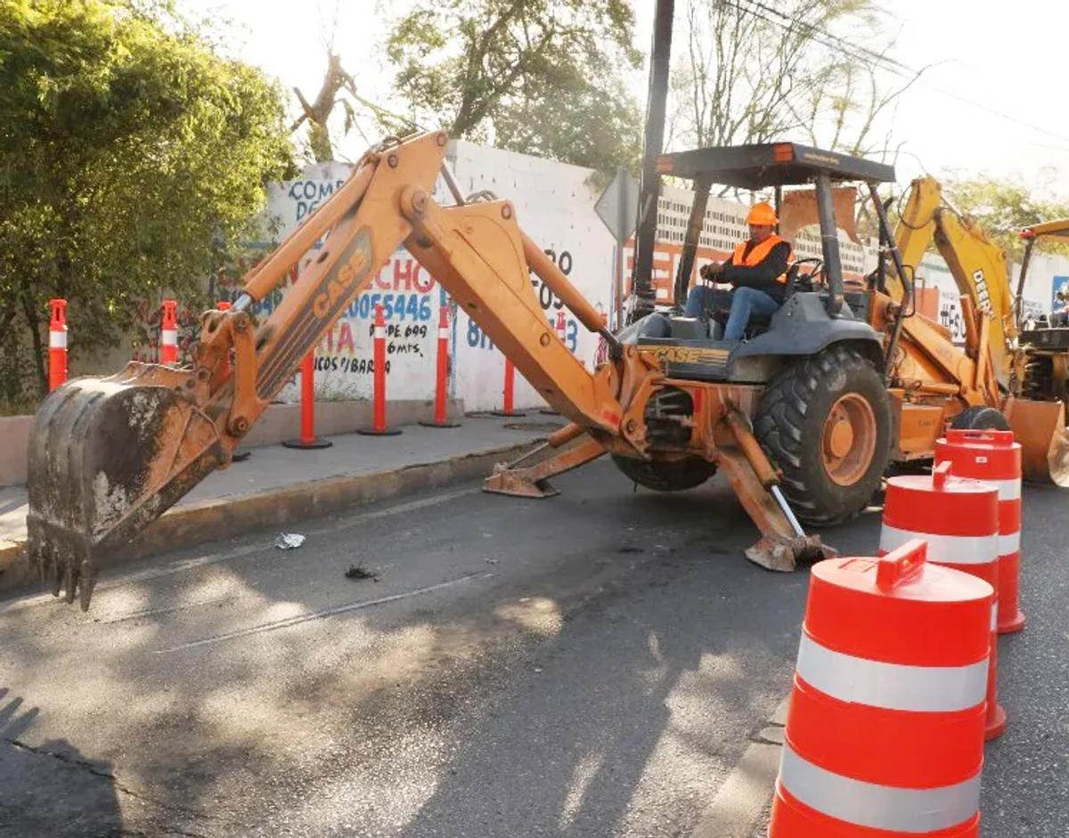 Retroexcavadora trabajando en una calle con trafitambos naranja con bandas blancas al frente y canalizadores en la banqueta