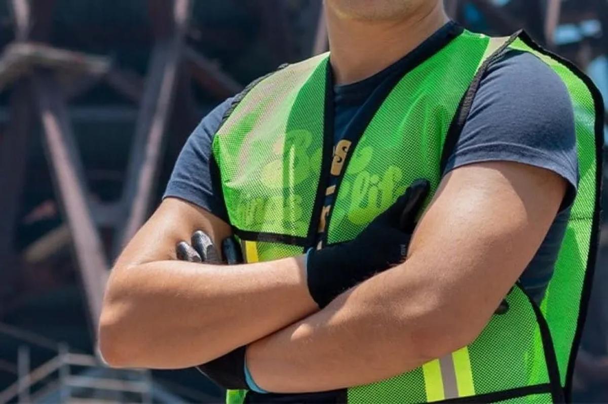 Trabajador de brazos cruzados con chaleco de malla verde fluorescente, bandas reflejantes y guantes negros