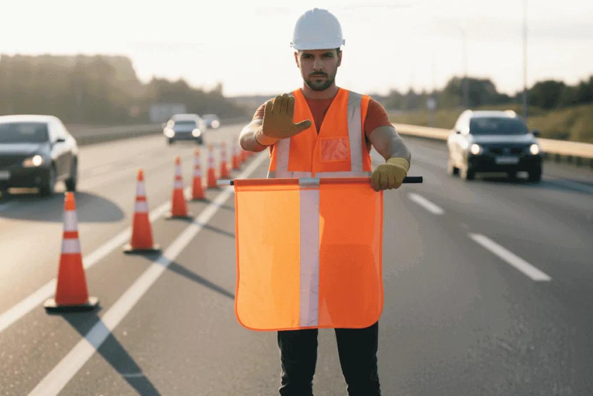 Banderero de frente con la palma en alto y el banderín naranja horizontal, señal de alto, en una carretera con conos