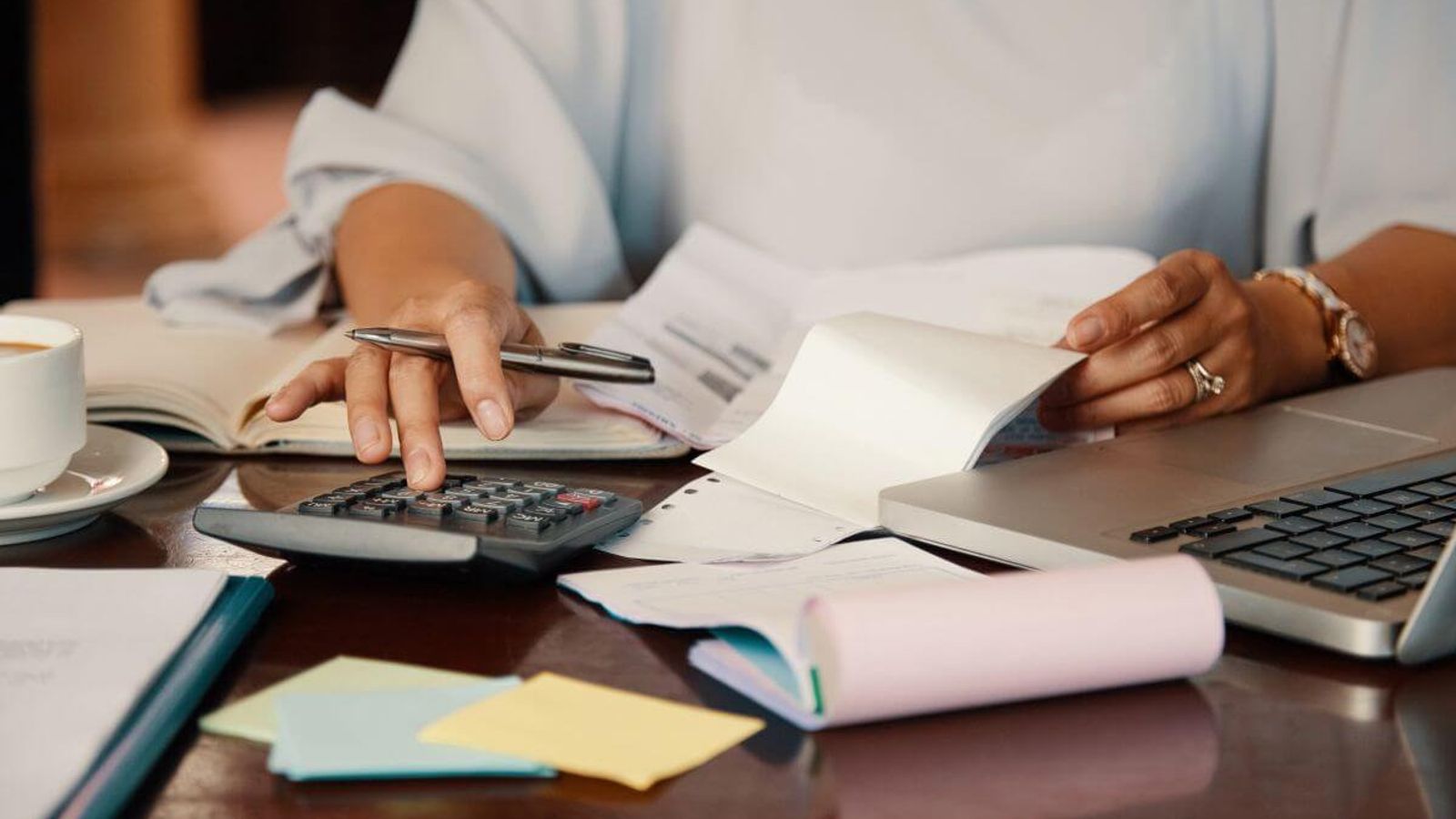 Mujer revisa recibos con una calculadora y una pluma en un escritorio con laptop, libreta y taza de café