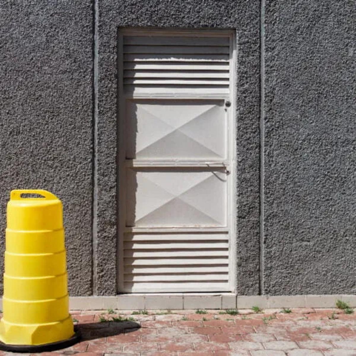 Trafitambo amarillo sin reflejante sobre base negra junto a un muro gris con puerta metálica blanca