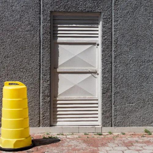 Trafitambo amarillo sin reflejante sobre base negra junto a un muro gris con puerta metálica blanca