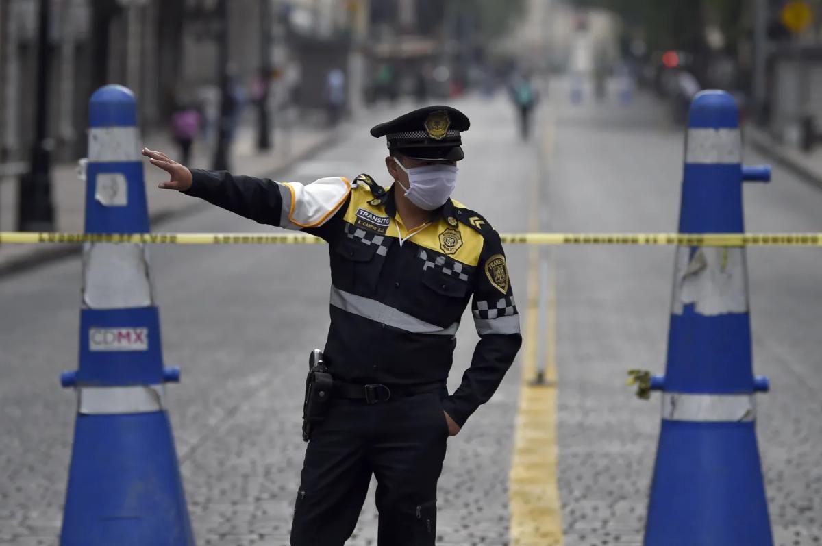 Policía de tránsito con cubrebocas da indicaciones detrás de una cinta amarilla, entre dos conos azules altos en una calle