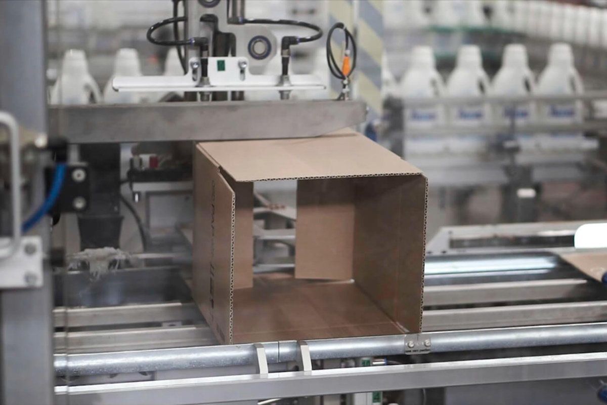 Máquina formadora de cajas con una caja de cartón abierta en la línea y botellas de plástico blancas al fondo