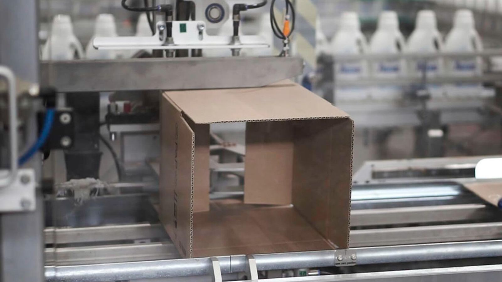 Máquina formadora de cajas con una caja de cartón abierta en la línea y botellas de plástico blancas al fondo