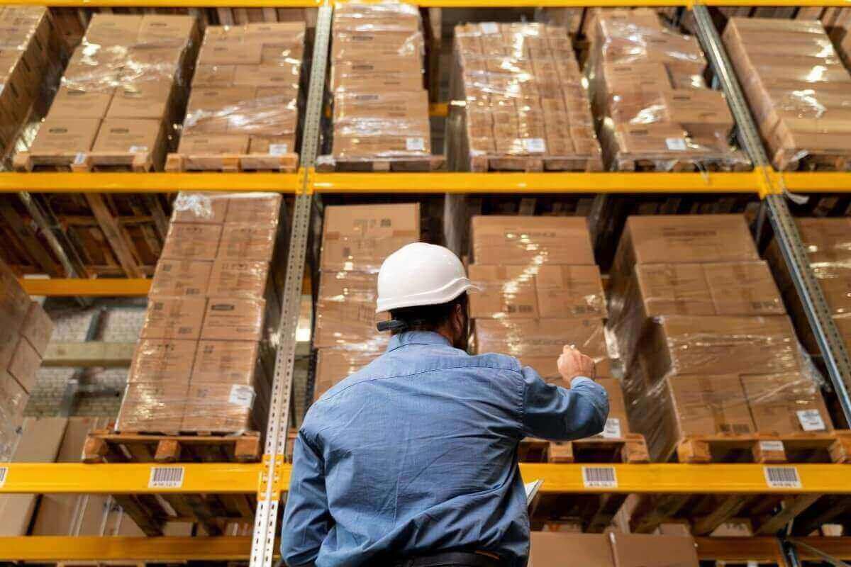Trabajador con casco blanco revisa tarimas de cajas emplayadas en racks de un almacén