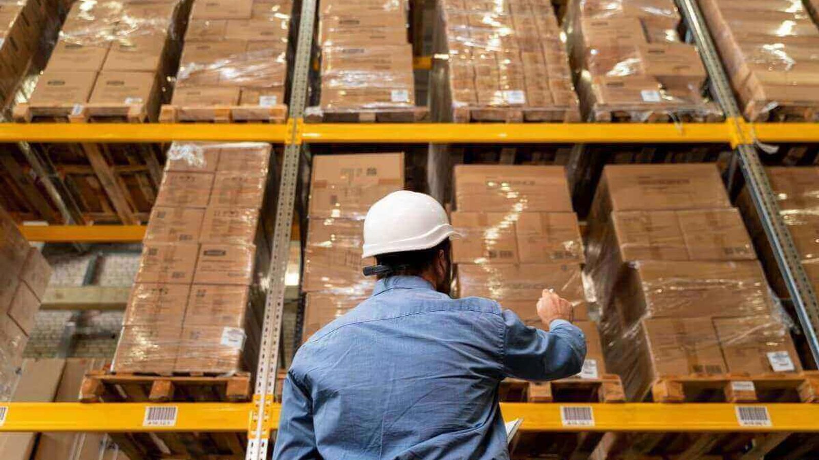 Trabajador con casco blanco revisa tarimas de cajas emplayadas en racks de un almacén