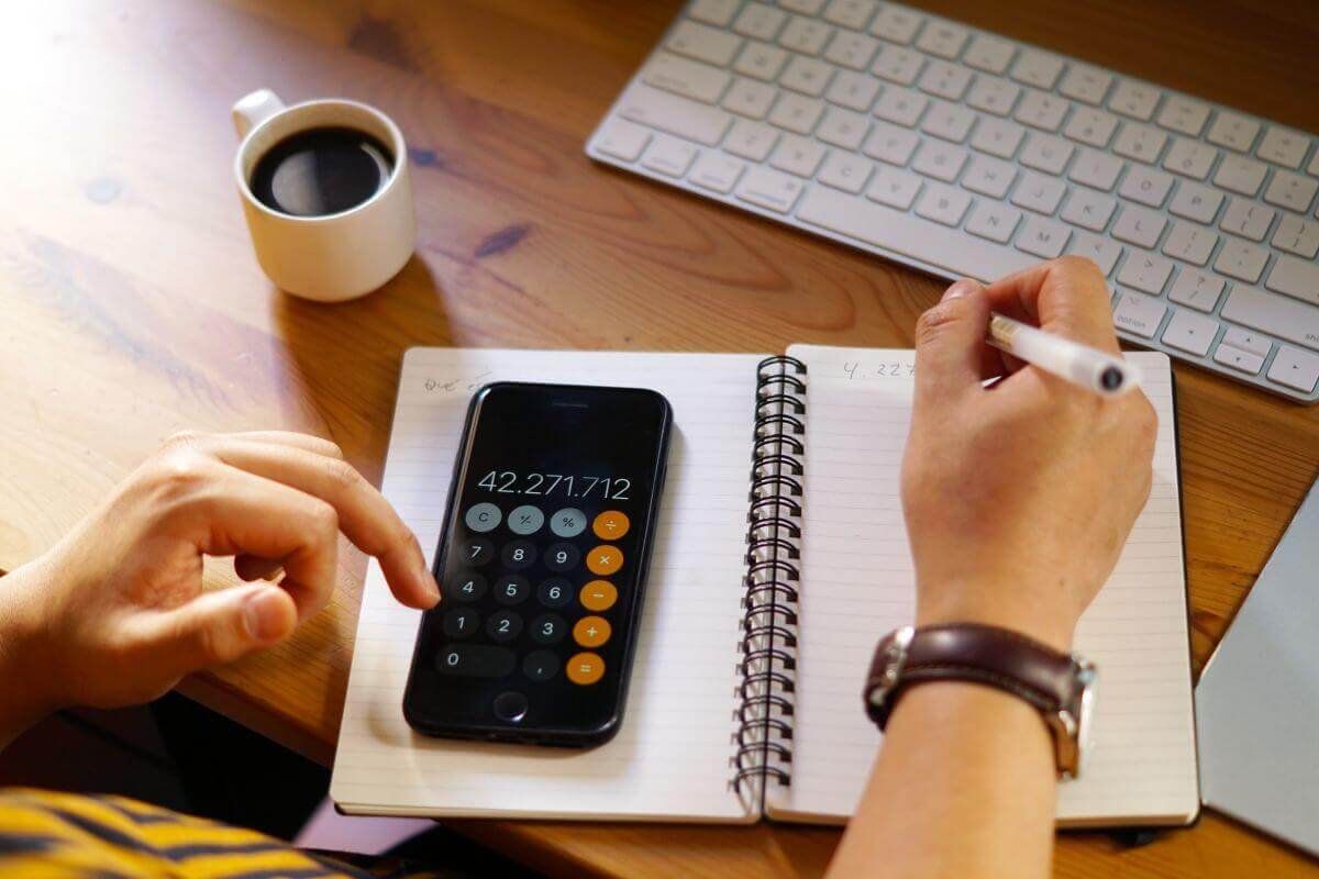 Persona usa la calculadora del celular sobre una libreta, junto a un teclado y una taza de café