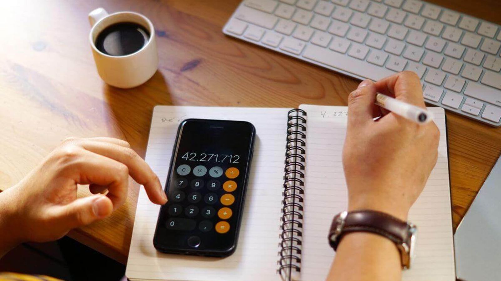 Persona usa la calculadora del celular sobre una libreta, junto a un teclado y una taza de café