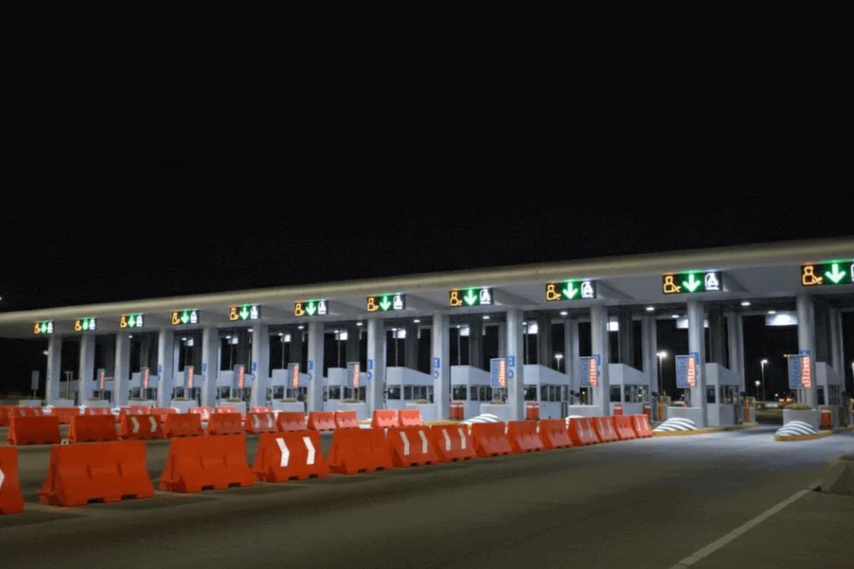 Caseta de cobro de noche con flechas verdes encendidas sobre los carriles y una fila de barreras viales naranjas al frente