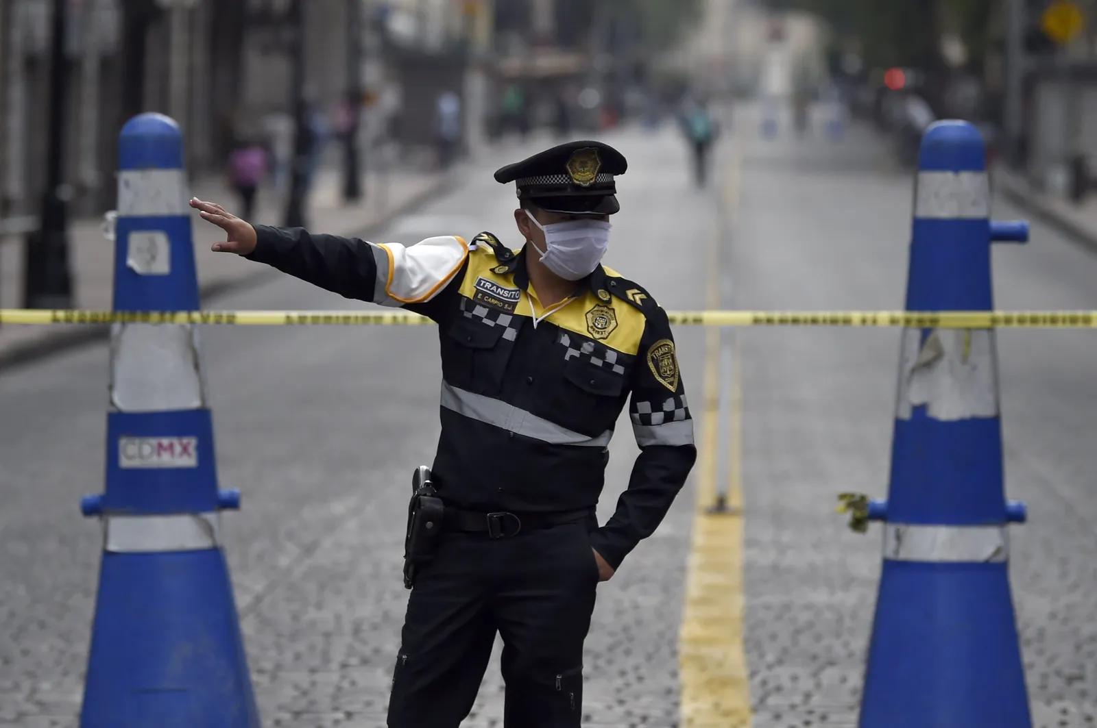 Policía de tránsito con cubrebocas da indicaciones detrás de una cinta amarilla, entre dos conos azules altos en una calle