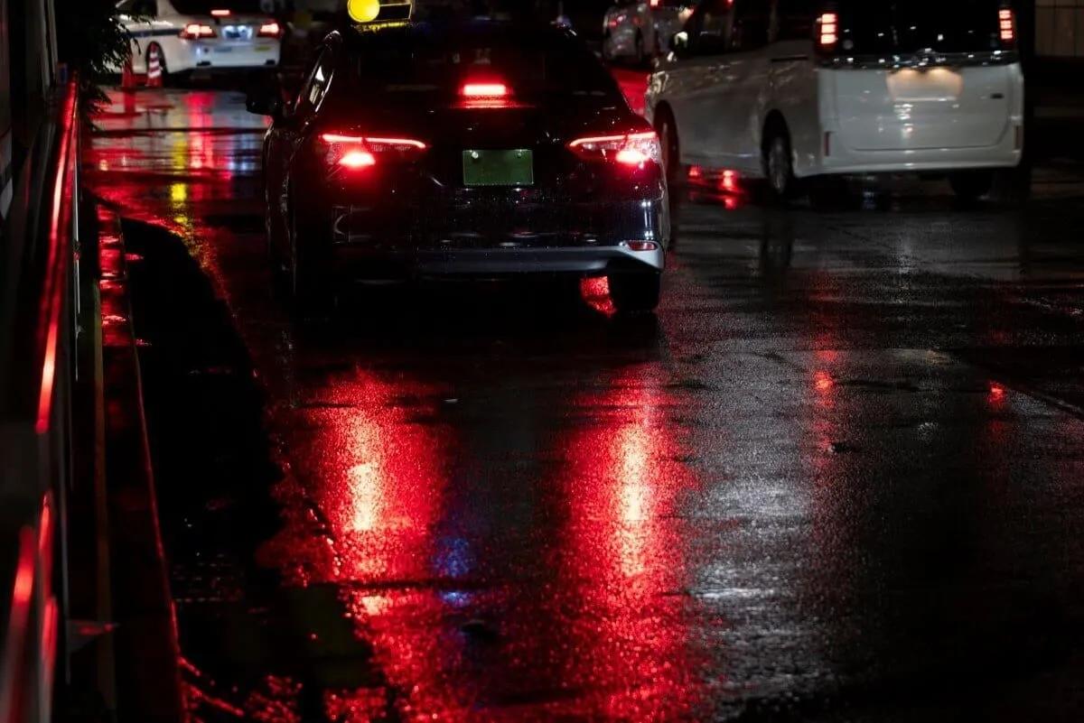 Autos con luces traseras rojas reflejadas en una calle mojada de noche, junto a una camioneta blanca