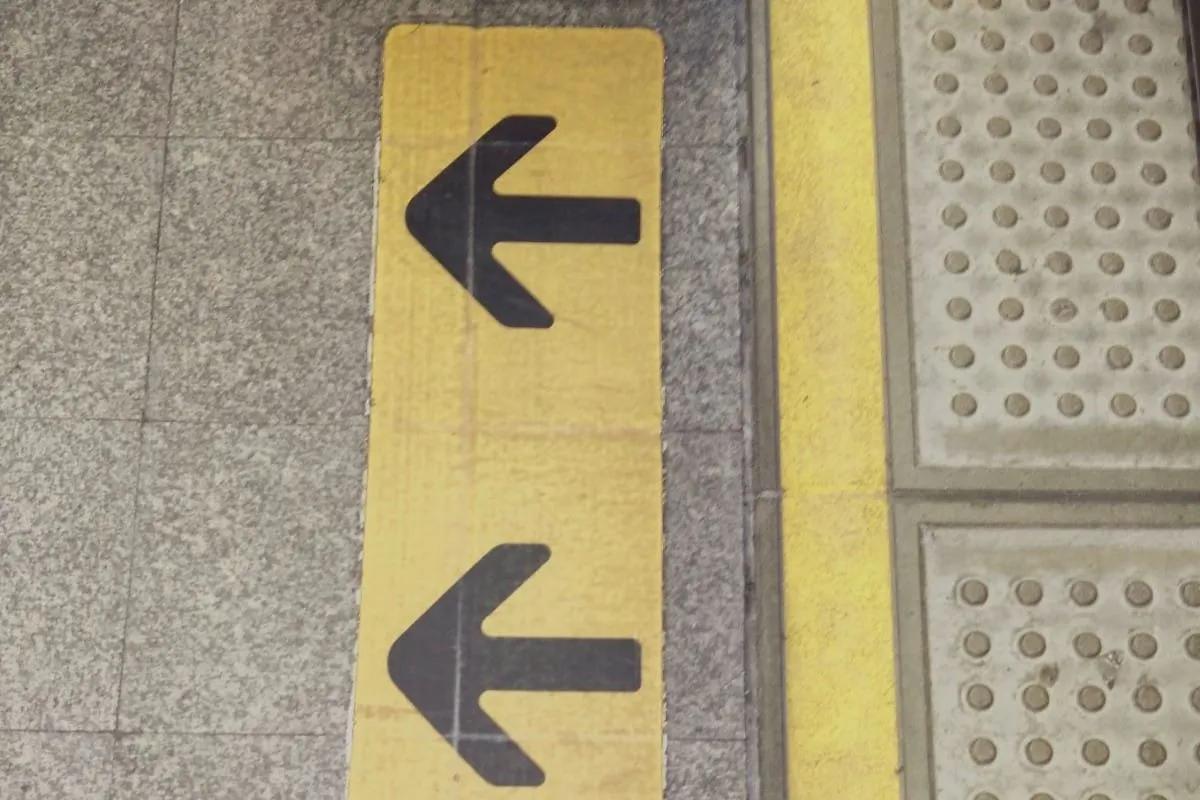 Franja amarilla con dos flechas negras junto a losetas podotáctiles grises de botones en el piso de una estación