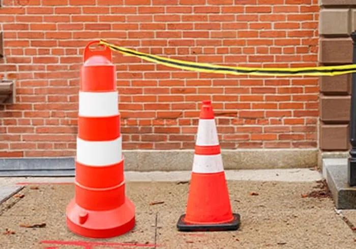 Trafitambo naranja con bandas blancas y cono vial unidos con cinta de precaución frente a un muro de ladrillo