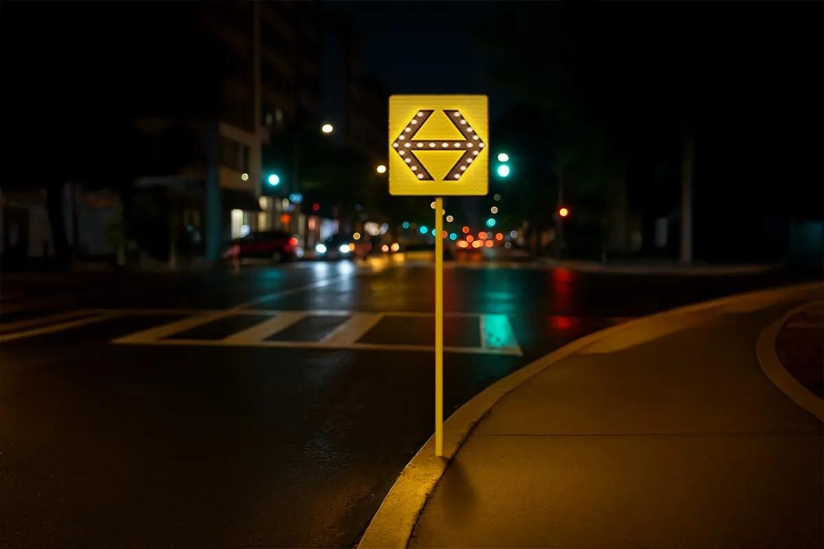 Señal vial cuadrada amarilla con una flecha doble iluminada por LED en una esquina urbana de noche