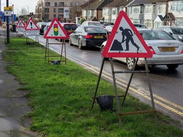 Señales de tránsito triangulares de obras, semáforo y cruce peatonal sobre caballetes en un camellón junto a una avenida