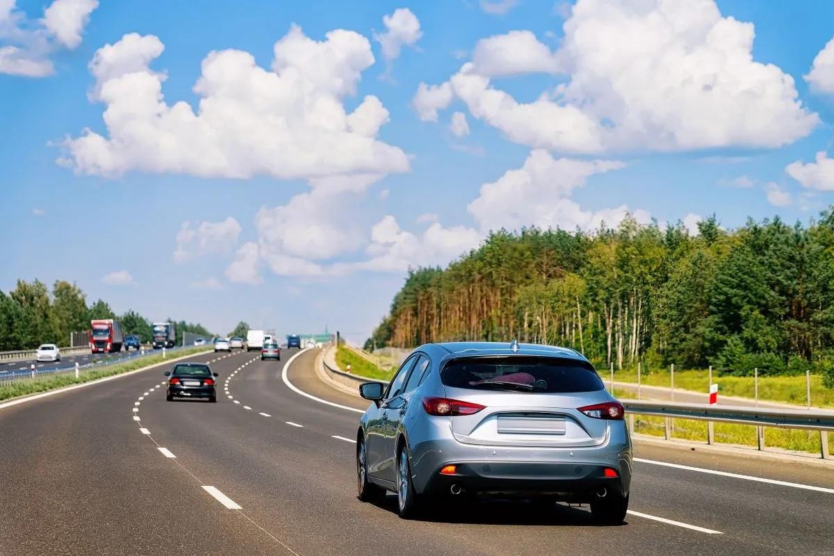Auto plateado circulando por una autopista de varios carriles entre bosque y cielo con nubes