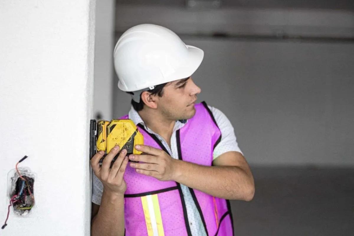 Trabajador con casco blanco y chaleco rosa con reflejante sostiene una herramienta amarilla contra un muro con cables