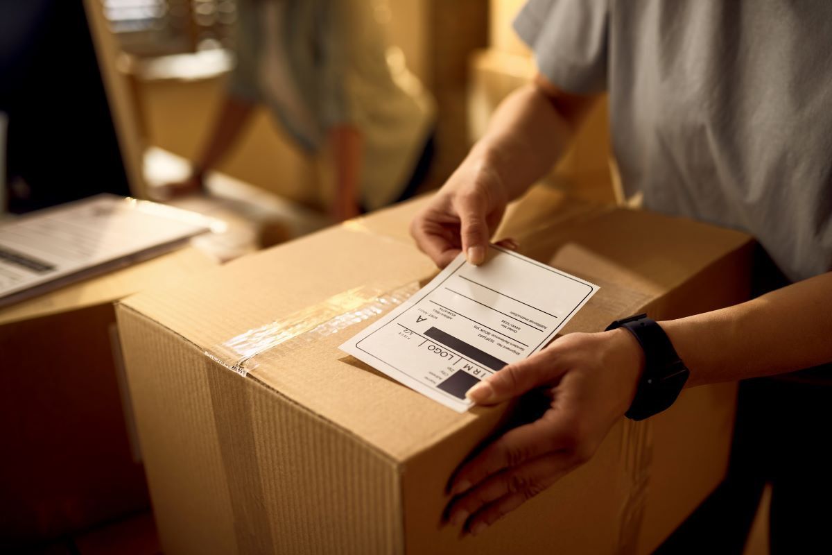 Persona pega una etiqueta de envío sobre una caja de cartón cerrada con cinta transparente, con más cajas al fondo