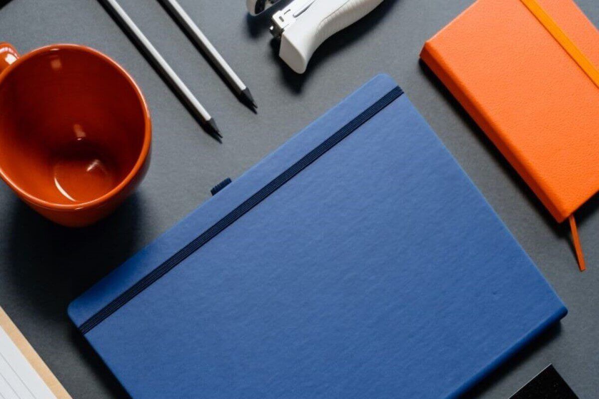 Libreta azul de pasta dura con elástico sobre un escritorio gris, junto a una libreta naranja, lápices y una taza