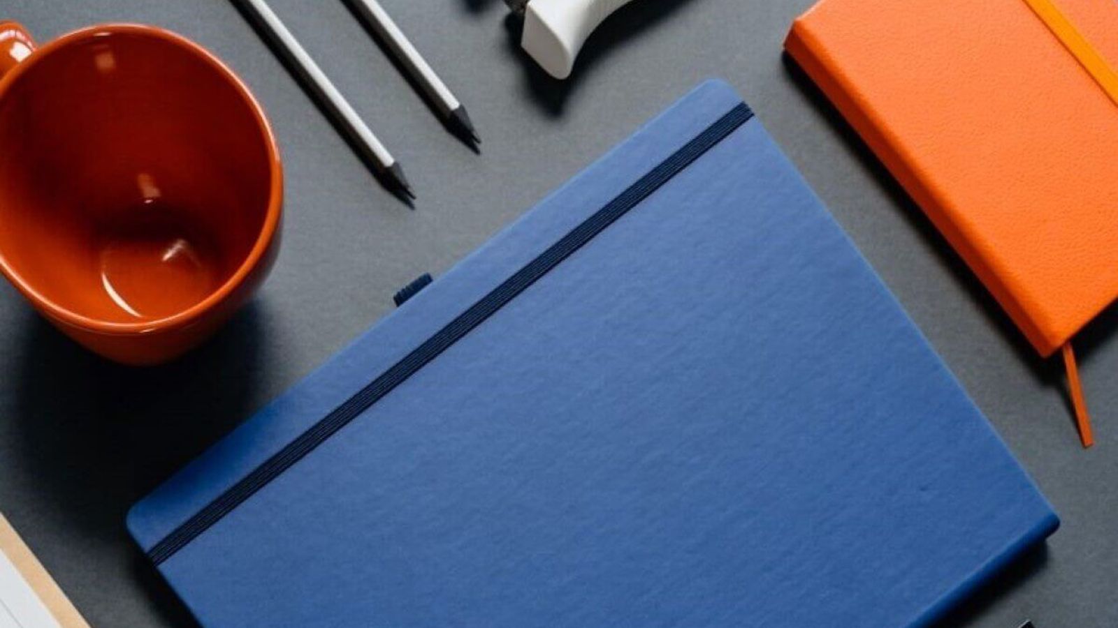 Libreta azul de pasta dura con elástico sobre un escritorio gris, junto a una libreta naranja, lápices y una taza